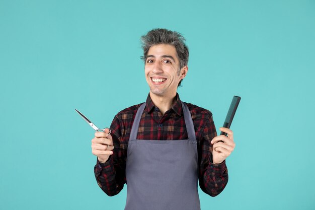 Vista superior de um jovem cabeleireiro feliz usando avental cinza e segurando o pente de tesoura na superfície de cor azul pastel