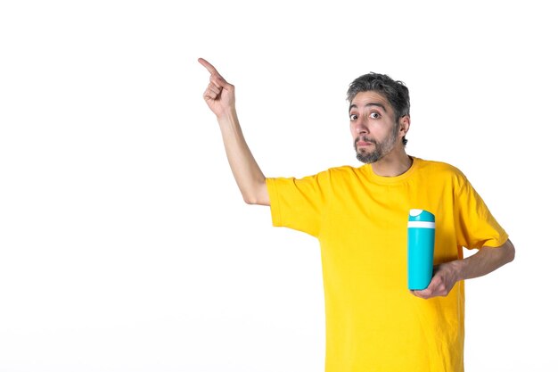 Vista superior de um jovem chocado com uma camisa amarela e uma garrafa térmica azul apontando para cima na superfície branca