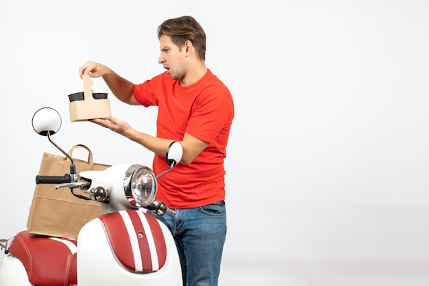 Vista superior de um jovem entregador confuso de uniforme vermelho em pé perto de scooter, olhando para o pedido na parede branca