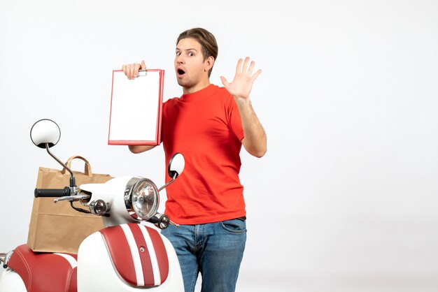 Vista superior de um jovem entregador emocionalmente confuso, de uniforme vermelho, em pé perto de uma scooter, mostrando o documento na parede branca