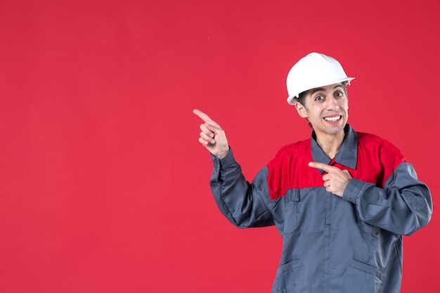 Vista superior de um jovem trabalhador curioso de uniforme com capacete e apontando para cima no lado direito na parede vermelha isolada