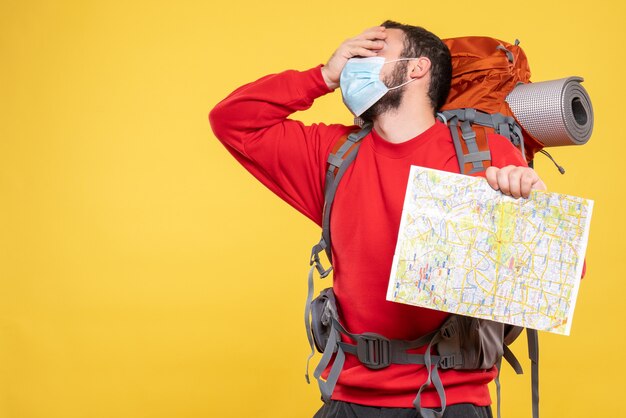 Vista superior de um jovem viajante exausto usando uma máscara médica com uma mochila segurando um mapa em amarelo