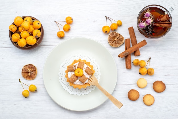 Vista superior de um pequeno bolo delicioso junto com coookies de canela e cerejas amarelas e chá na luz, biscoito de bolacha