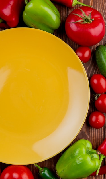 Vista superior de um prato amarelo vazio e legumes frescos coloridos pimentões tomates e pepinos em madeira rústica