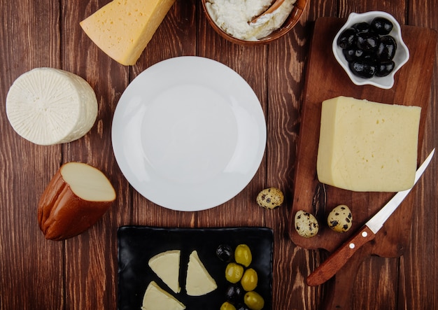Vista superior de um prato branco vazio e diferentes tipos de queijo com azeitonas em conserva e ovos de codorna, dispostas na mesa rústica