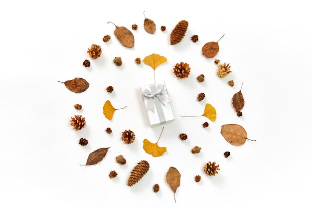 Vista superior de um presente no meio de uma coroa de flores feita de folhas de outono e cones de coníferas em branco