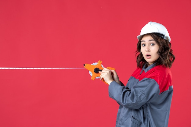 Vista superior de uma arquiteta chocada de uniforme, com capacete abrindo a fita métrica em fundo vermelho isolado