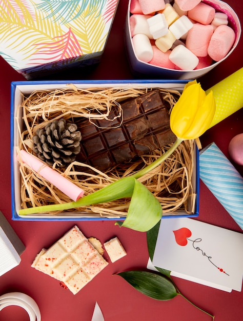 Vista superior de uma caixa de presente aberta com flor de tulipa de cor amarela, barra de chocolate escura, cone e palha e uma caixa em forma de coração cheia de marshmallow na mesa vermelha escura
