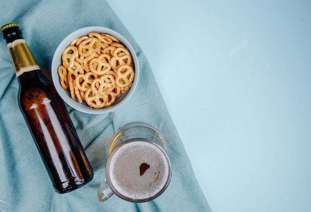 Vista superior de uma caneca de cerveja e uma garrafa com mini pretzels em uma tigela em azul com espaço de cópia
