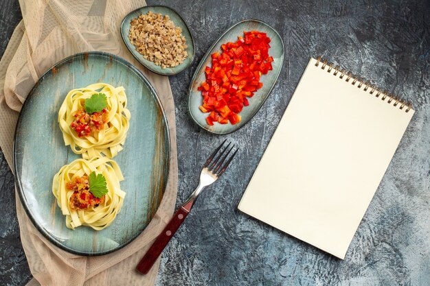 Vista superior de uma deliciosa refeição de massa em um prato azul servido com tomate e carne para o jantar em uma toalha de cor bronzeada seus ingredientes ao lado de um caderno fechado