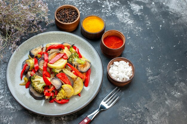 Vista superior de uma deliciosa refeição vegetariana em um prato e um garfo ao lado de diferentes especiarias no lado direito em fundo escuro