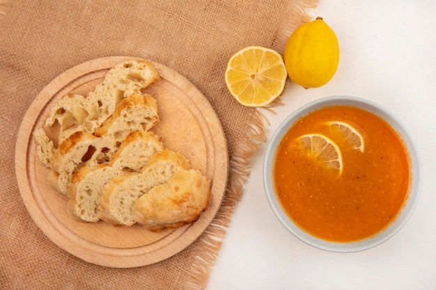 Vista superior de uma deliciosa sopa de lentilhas em uma tigela com fatias de pão em uma placa de cozinha de madeira em um pano de saco com limões em uma superfície branca