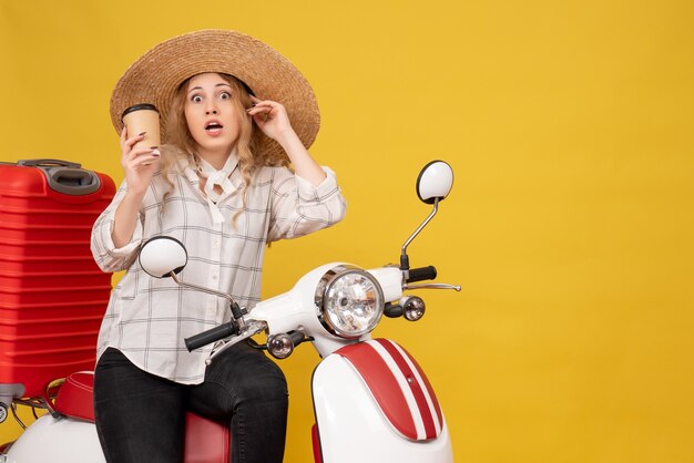 Vista superior de uma jovem chocada com um chapéu, sentada na motocicleta e segurando café