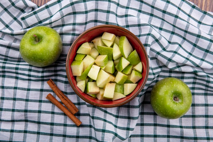 Foto grátis vista superior de uma maçã fresca inteira com fatias de maçã picada em uma tigela vermelha com paus de canela em uma toalha de mesa quadriculada