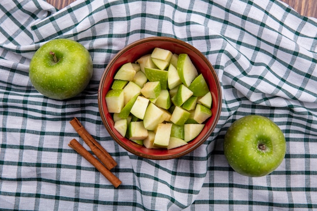 Vista superior de uma maçã fresca inteira com fatias de maçã picada em uma tigela vermelha com paus de canela em uma toalha de mesa quadriculada
