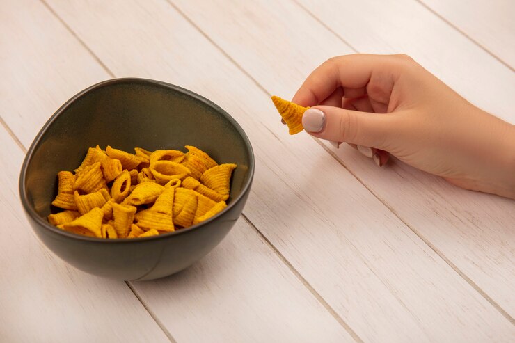 Foto grátis vista superior de uma mão feminina segurando salgadinhos de milho em forma de cone com uma tigela de batatas fritas em uma mesa de madeira bege
