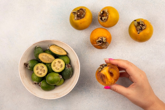 Vista superior de uma mão feminina segurando uma fruta madura de meio caqui com feijoas em uma tigela e caquis isolados em uma superfície cinza