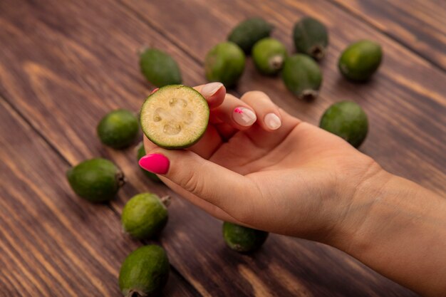 Vista superior de uma mão feminina segurando uma meia feijoa fresca com feijoas isoladas em uma superfície de madeira