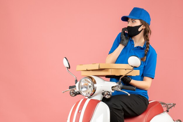 Vista superior de uma mensageira assustada usando máscara médica e luvas, sentada na scooter, entregando pedidos em fundo cor de pêssego