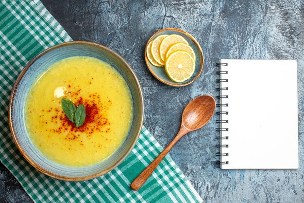 Vista superior de uma panela azul com sopa saborosa servida com hortelã e pimenta ao lado do caderno espiral de colher de pau de limão picado sobre fundo azul