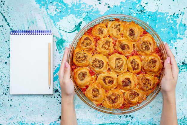 Vista superior de uma refeição de massa cozida com carne picada e molho de tomate dentro de uma panela de vidro com um bloco de notas em azul brilhante, cozinhando massa de carne para assar comida