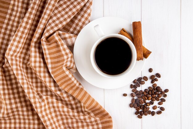 Vista superior de uma xícara de café com paus de canela e grãos de café espalhados em fundo branco de madeira