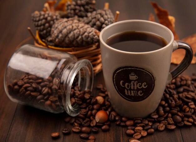 Vista superior de uma xícara de café com pinhas em um balde com grãos de café caindo de uma jarra de vidro sobre uma superfície de madeira