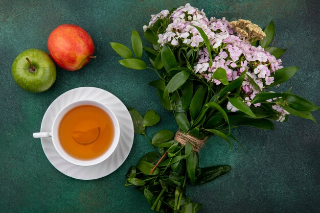 Vista superior de uma xícara de chá com maçãs e um buquê de flores em uma superfície verde