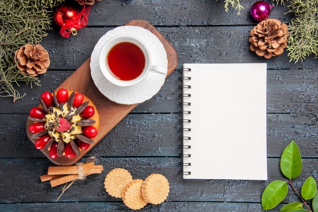 Vista superior de uma xícara de chá e bolo de frutas vermelhas em folhas de brinquedos de Natal de pinhas prato de servir de madeira e um caderno na mesa de madeira escura