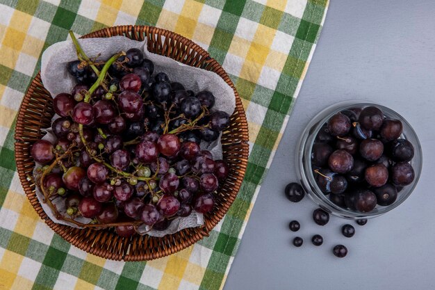 Vista superior de uva preta em uma cesta em tecido xadrez e bagas de uva em uma tigela sobre fundo cinza