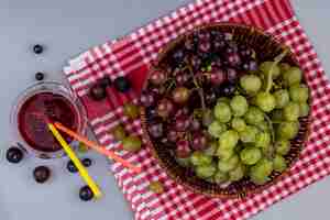 Foto grátis vista superior de uvas em uma cesta em pano xadrez e copo de suco de uva em fundo cinza