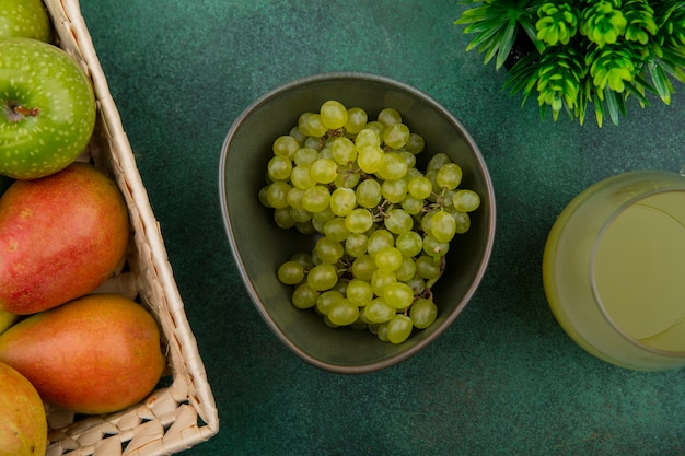 Vista superior de uvas verdes em uma tigela com uma maçã verde e peras em uma cesta com suco em um fundo verde