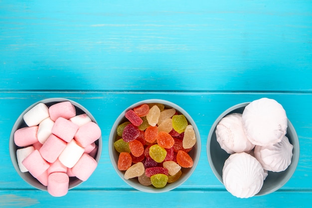 Vista superior de vários doces de marmelada colorida de doces com zéfiro branco e marshmallows em tigelas em azul