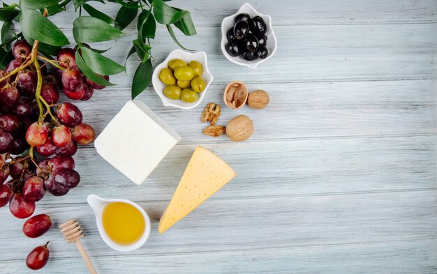 Vista superior de vários tipos de queijo com uvas frescas, nozes, mel e azeitonas em conserva na mesa de madeira cinza com espaço de cópia