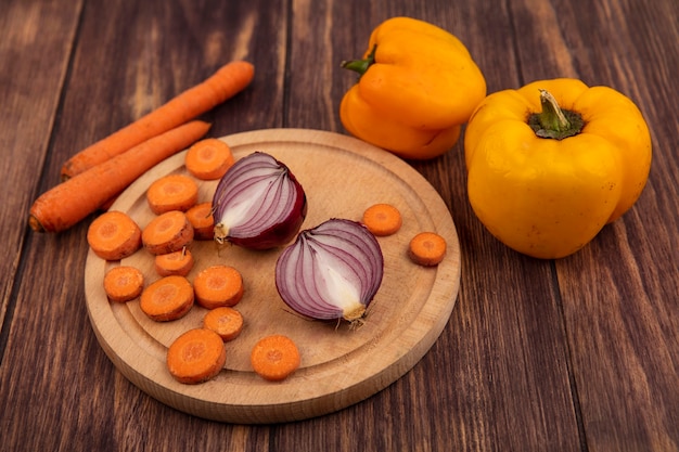 Vista superior de vegetais frescos, como cenouras picadas e cebolas vermelhas em uma placa de cozinha de madeira com cenouras e pimentões amarelos isolados em um fundo de madeira