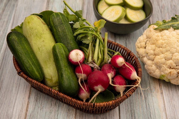 Vista superior de vegetais orgânicos, como abobrinhas de pepino e rabanetes em um balde com couve-flor isolada em um fundo cinza de madeira