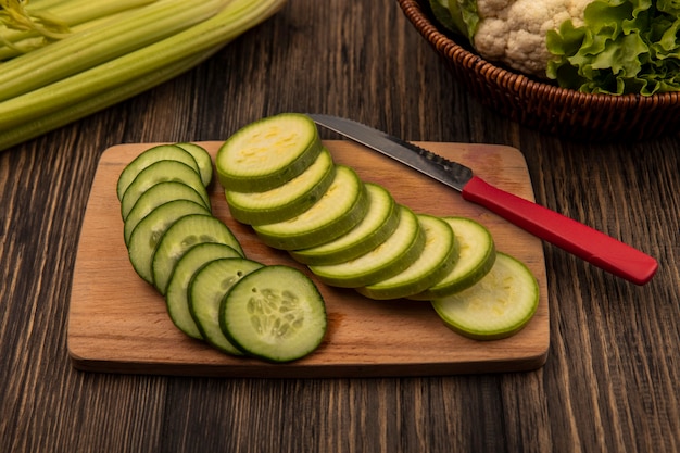 Vista superior de vegetais picados como pepino e abobrinha em uma placa de cozinha de madeira com faca com couve-flor e alface em um balde com aipo isolado em uma parede de madeira