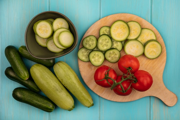 Vista superior de vegetais saudáveis, como tomates picados, pepinos e abobrinhas em uma placa de cozinha de madeira com pepinos e abobrinhas isolados em uma superfície de madeira azul