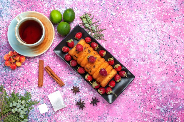 Vista superior delicioso bolo assado dentro da fôrma preta com chá de morangos vermelhos frescos e limões na mesa rosa.