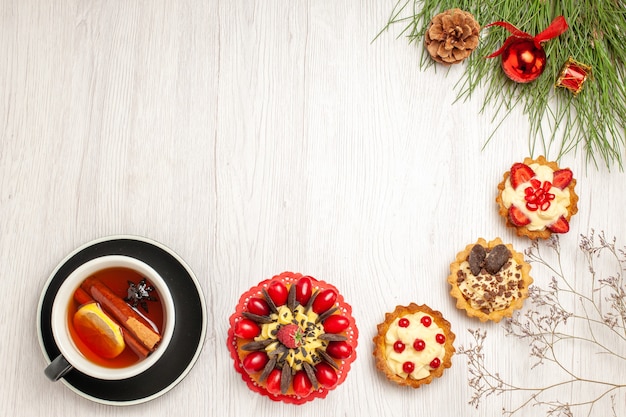 Vista superior distante uma xícara de tortas de bolo de baga e chá de canela e limão na parte inferior e as folhas do pinheiro com brinquedos de natal no canto superior direito do chão de madeira branco