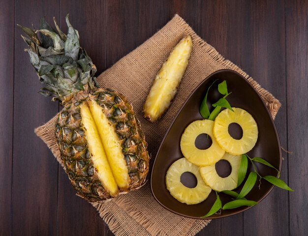 Vista superior do abacaxi com um pedaço cortado de frutas inteiras e tigela de fatias de abacaxi no saco e na superfície de madeira