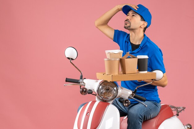 Vista superior do atencioso mensageiro usando chapéu, sentado na scooter em um pêssego pastel