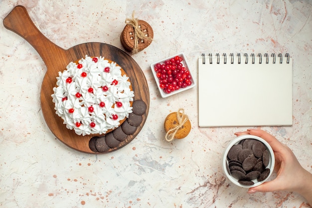 Vista superior do bolo com creme de confeiteiro branco na tigela de tábua com frutas tigela de chocolate em biscoitos de mão de mulher amarrados com corda na mesa cinza claro