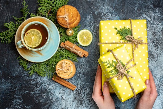 Vista superior do bolo delicioso cremoso e ramos de abeto, limão, canela, limão, mão segurando uma das caixas de presente amarelo, uma xícara de chá preto sobre fundo escuro