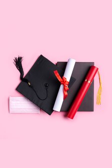 Vista superior do boné quadrado acadêmico de graduação com diploma de graduação e máscara isolada no fundo da mesa-de-rosa.