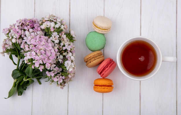 Vista superior do buquê de flores com uma xícara de chá e macarons coloridos em uma superfície branca