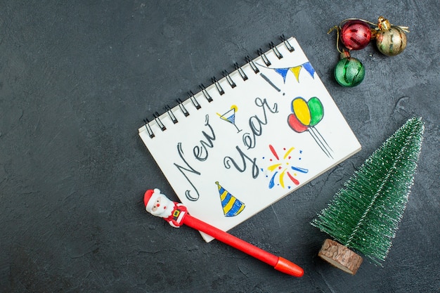 Vista superior do caderno espiral com a escrita de ano novo e uma caneta ao lado dos acessórios de decoração da árvore de natal em fundo escuro