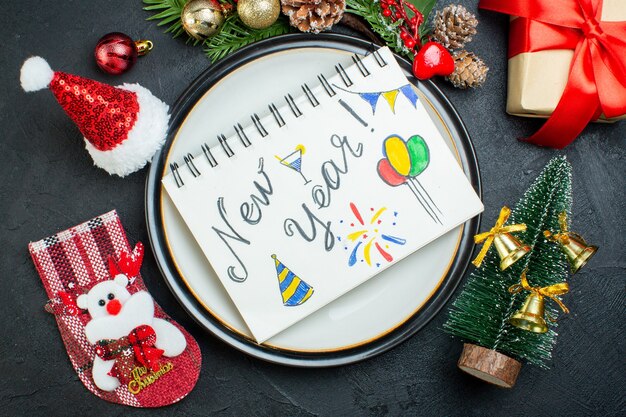 Vista superior do caderno espiral com caneta no prato de jantar árvore de natal ramos de coníferas cone caixa de presente chapéu de Papai Noel meia de Natal em fundo preto