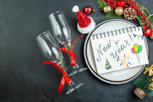 Vista superior do caderno espiral com caneta no prato de jantar árvore de natal ramos de coníferas cone caixa de presente chapéu de papai noel taças de vidro caídas no fundo preto