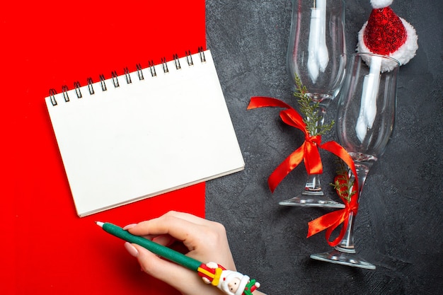 Vista superior do caderno espiral e a mão segurando uma caneta ao lado de taças de vidro, chapéu de Papai Noel em fundo vermelho e preto